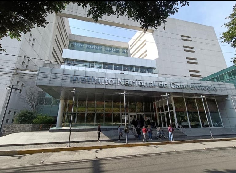 Modern institutional building with Instituto Nacional de Cancerología signage, glass storefront entrance, and several people standing outside.