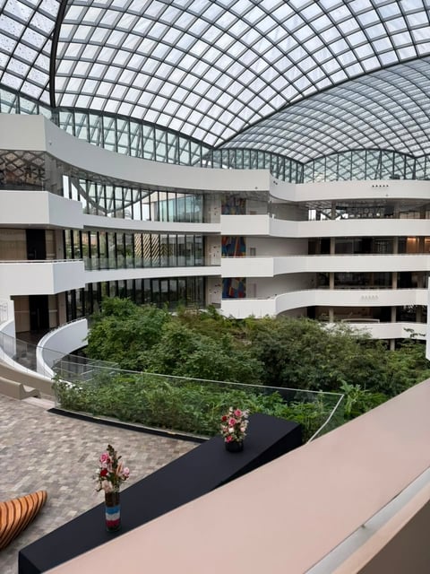Modern multi-level building atrium with curved white balconies, glass gridded ceiling, and central green landscaping with planted beds.
