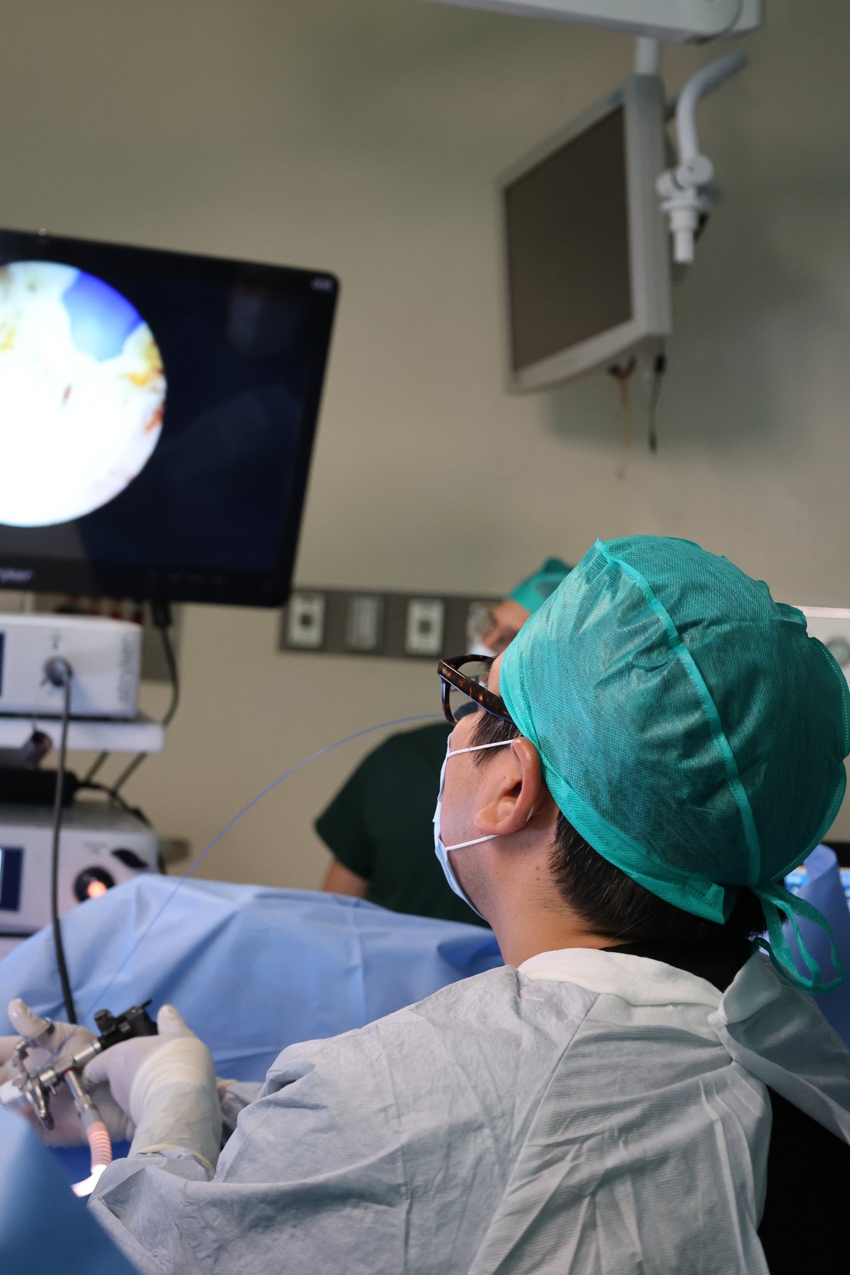 Surgeon in turquoise surgical cap and mask performing operation in medical facility with overhead lighting and monitor