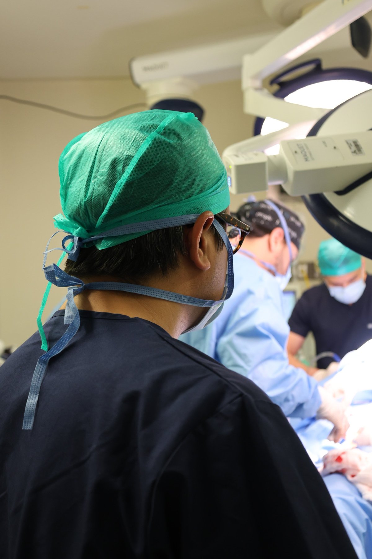 Surgeon wearing green scrub cap and protective eyewear in operating room with surgical equipment overhead