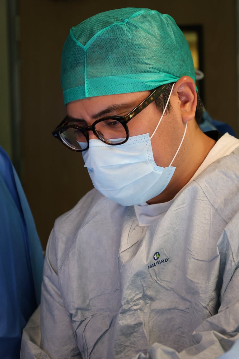 Medical professional wearing surgical attire with green cap, glasses, and face mask in clinical setting