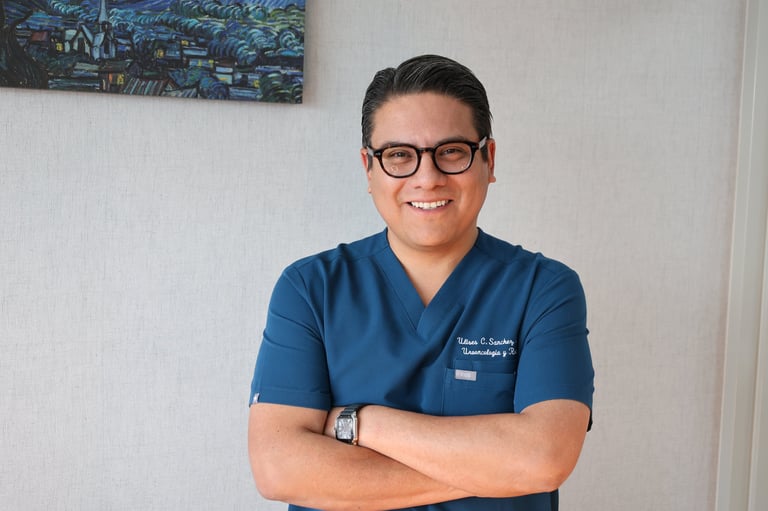 Man wearing blue scrubs and glasses stands with arms crossed, smiling at camera in medical office setting