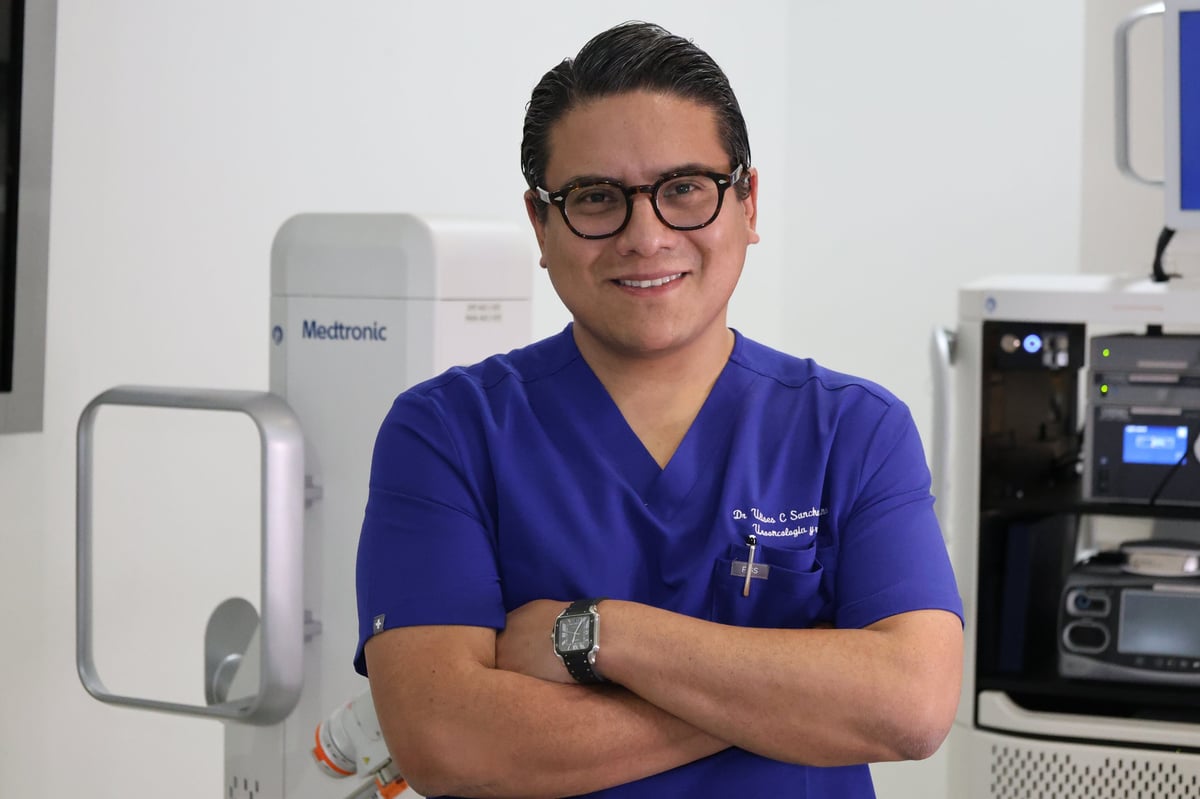 Male healthcare professional in blue scrubs and glasses standing with arms crossed in a medical office with equipment behind him