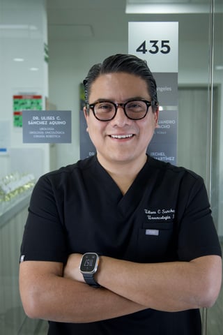 Male healthcare professional smiling at camera wearing glasses and black medical scrubs in an office setting
